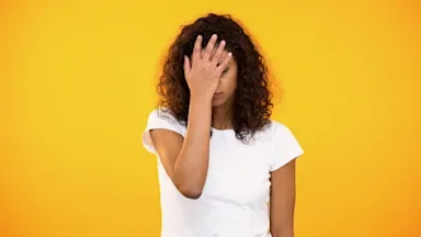 Portrait of a young woman with her hand on her forehead. She looks like she has a headache. She is standing on a yellow studio background.
Motortion/iStock via Getty Images