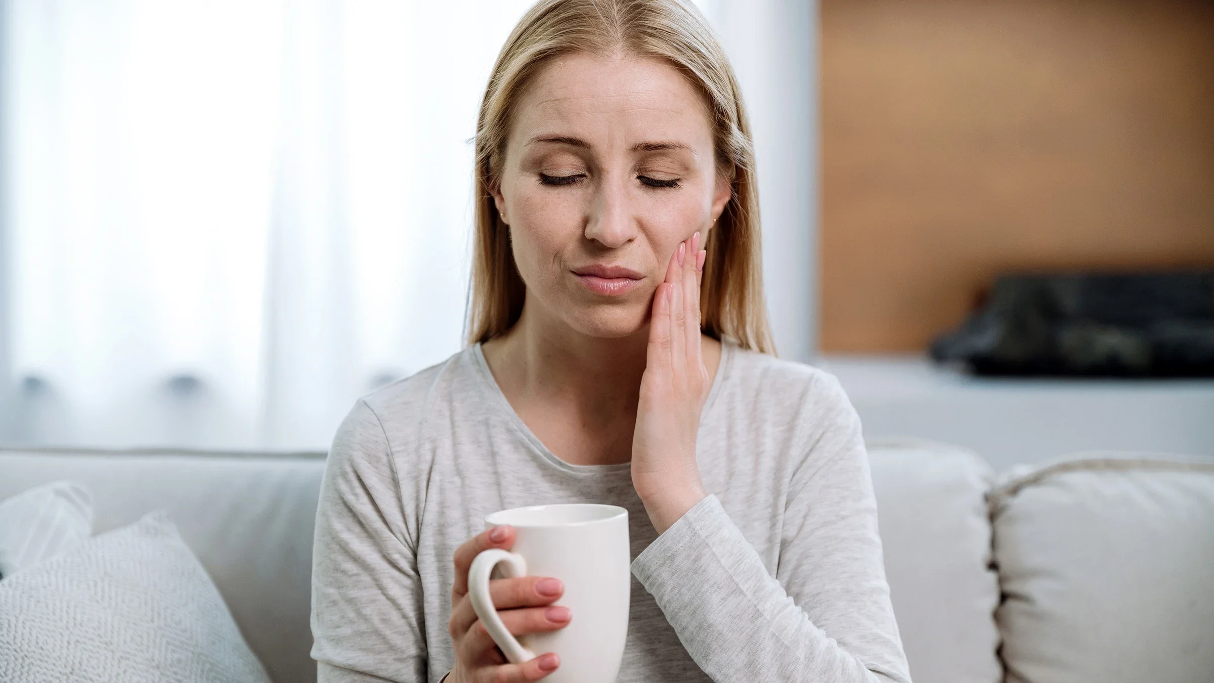 While holding a mug, a woman touches her cheek as she experiences tooth pain.
brizmaker/iStock via Getty Images Plus