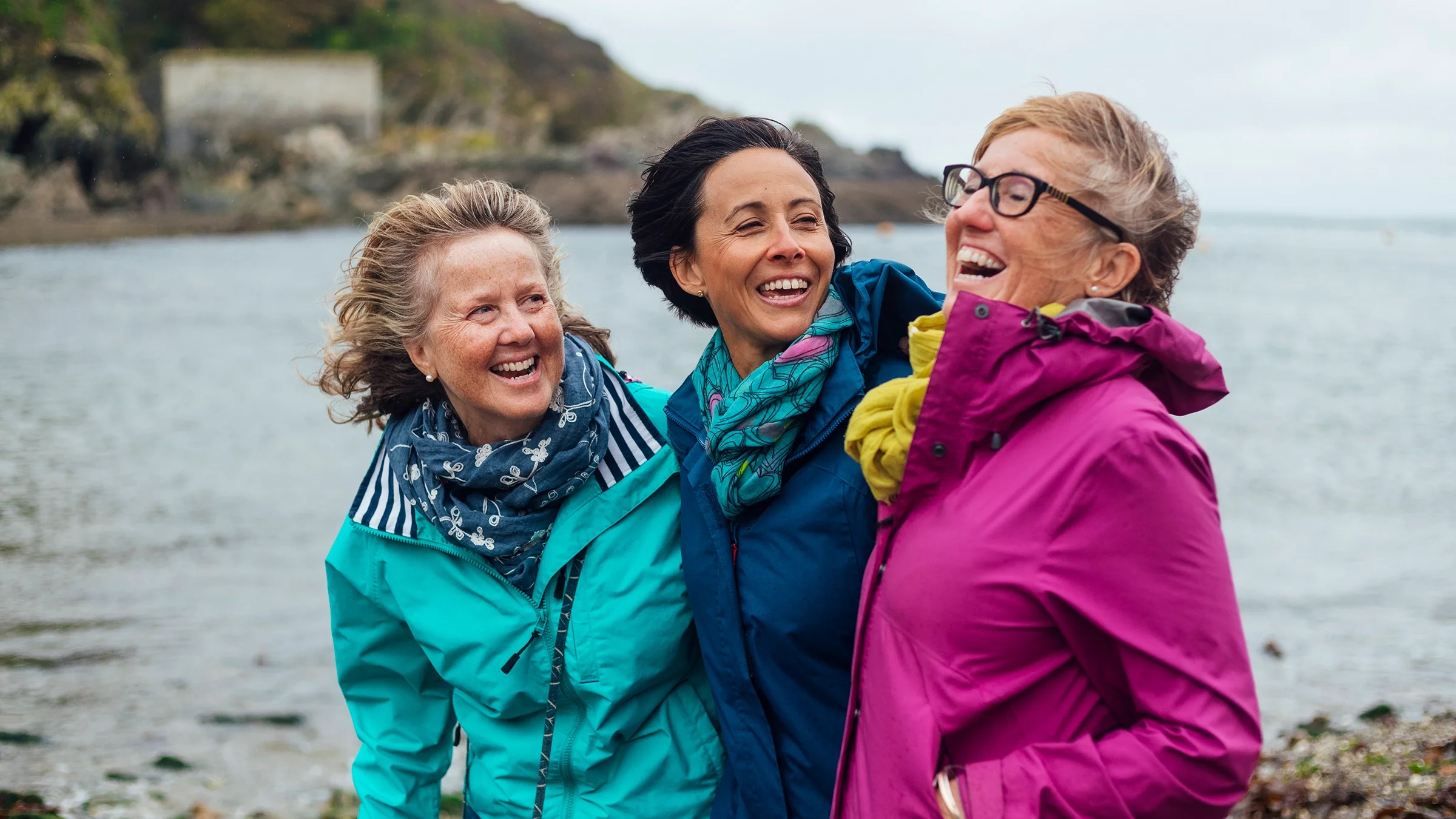 Three friends are wearing raincoats on a windy beach. They are laughing and smiling at one another.