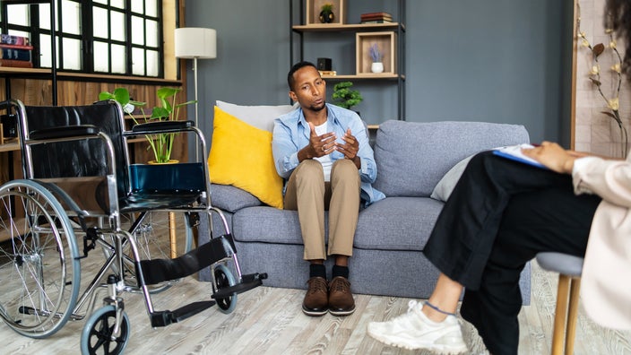 A man sits on a couch beside his empty wheelchair and talks to a therapist.