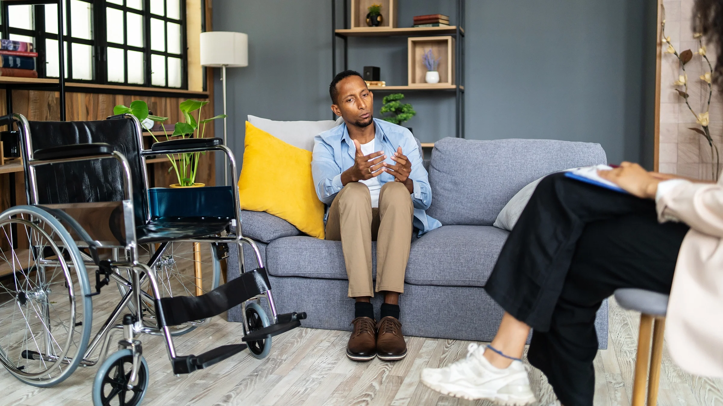 A man sits on a couch beside his empty wheelchair and talks to a therapist.