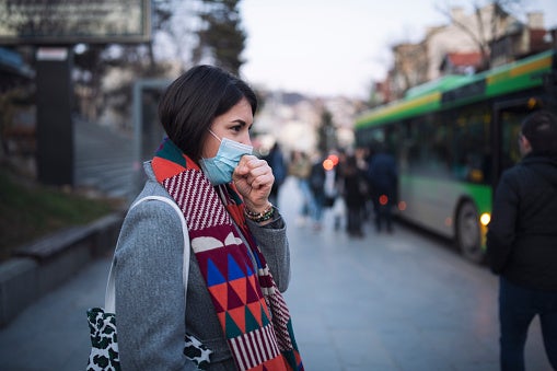 Woman standing outside at a bus stop wearing a blue medical face mask while making a coughing motion with her hand near her face.