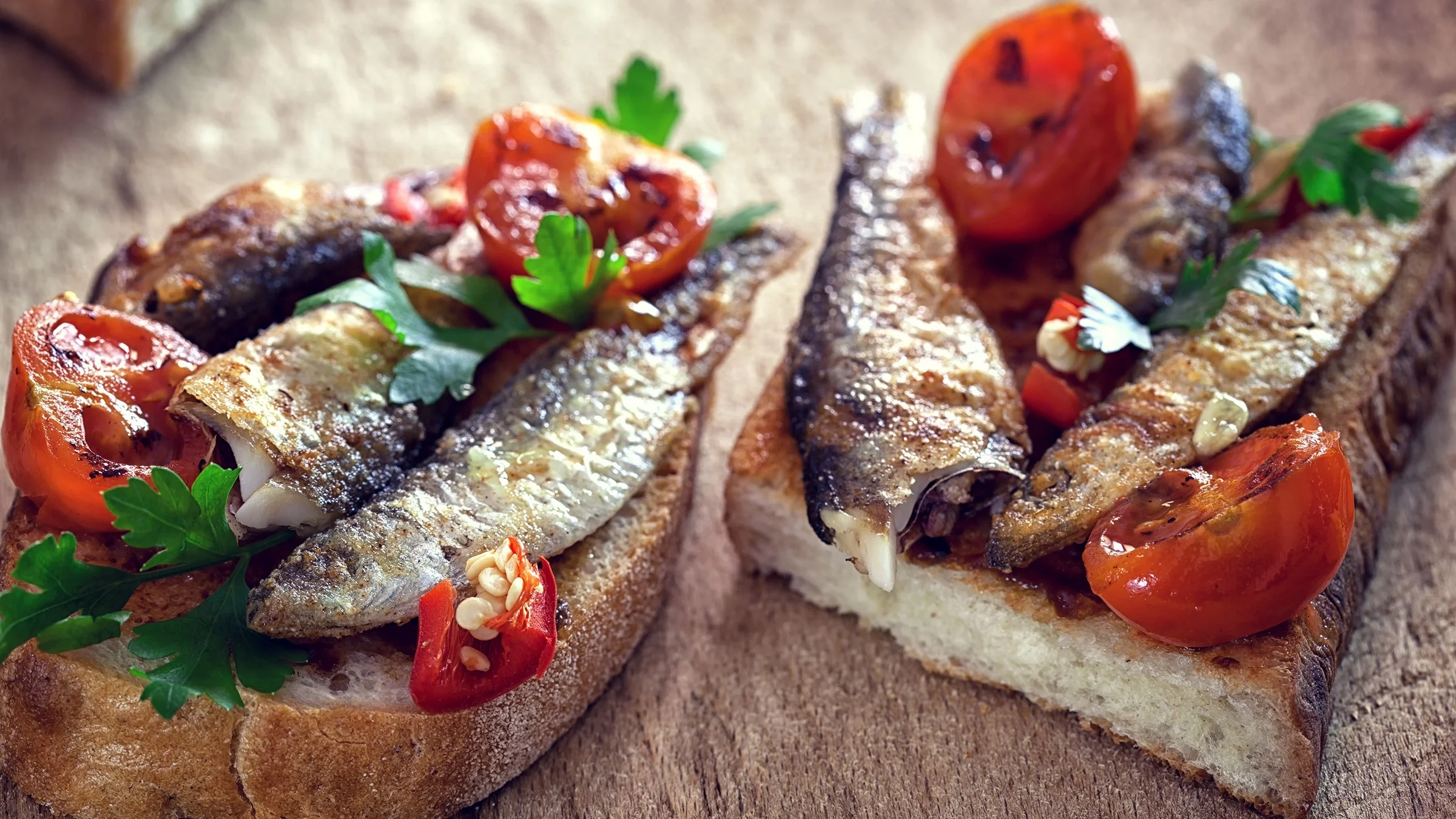 Fried sardines and tomatoes on toasted bread. All sitting on a wooden cutting board.