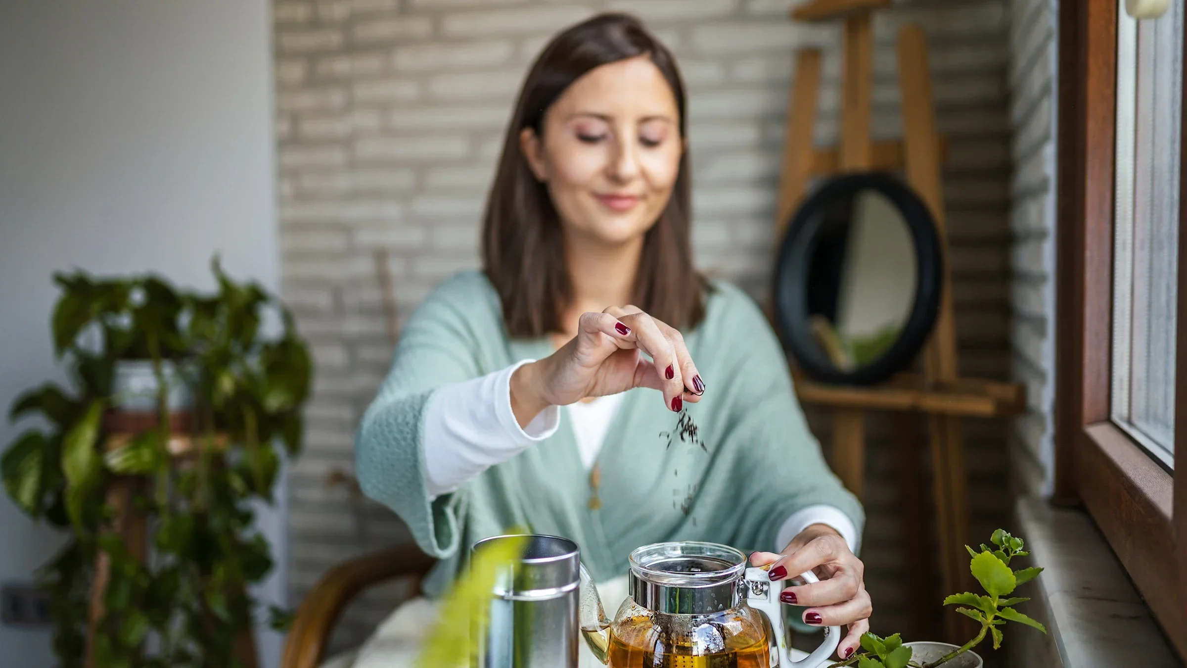 Woman sitting at her kitchen table making a large pot of green tea. She is sprinkling in more tea leaves into the glass tea pot.
Kemal Yildirim/E+ via Getty Images