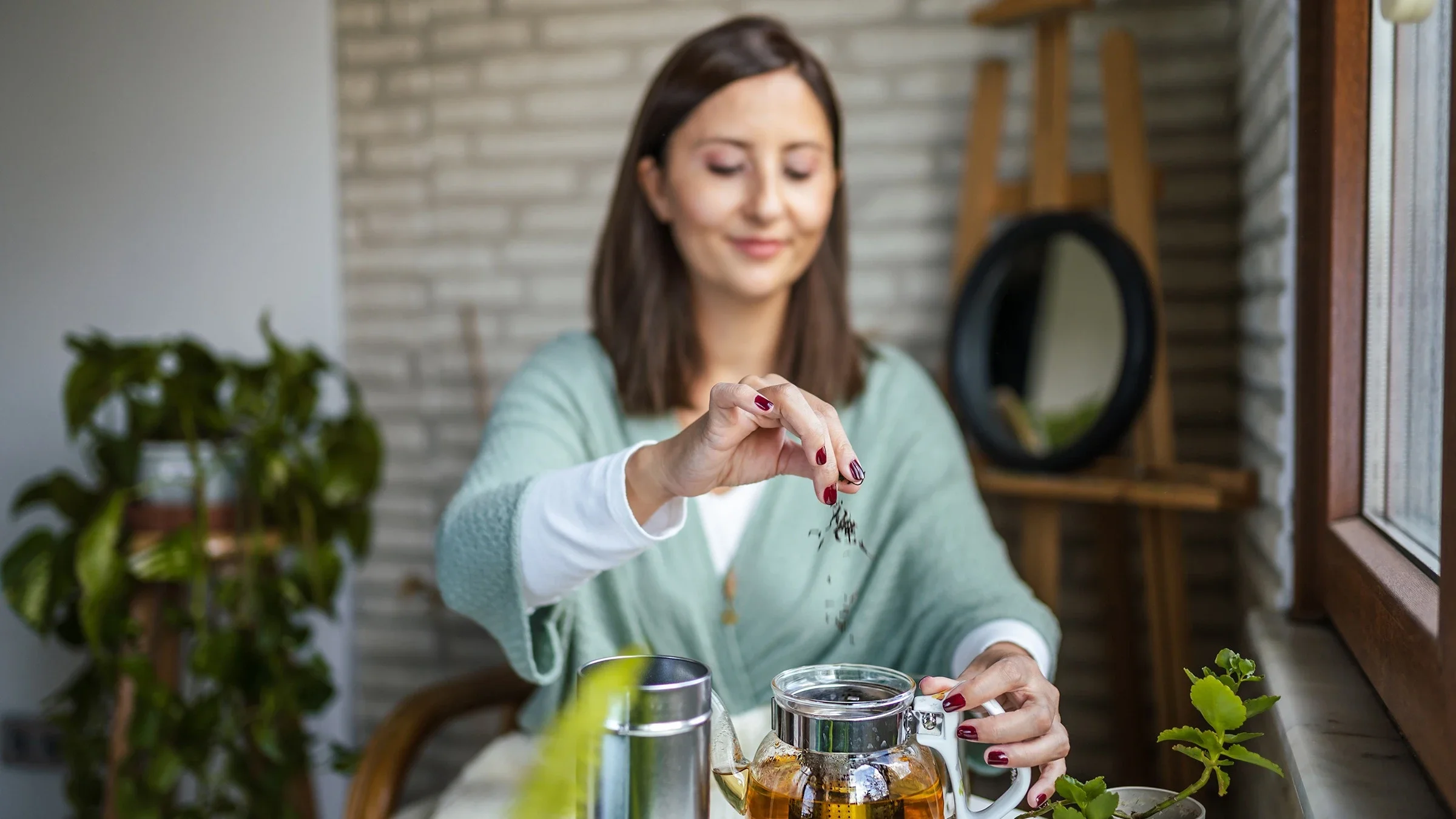 Woman sitting at her kitchen table making a large pot of green tea. She is sprinkling in more tea leaves into the glass tea pot.