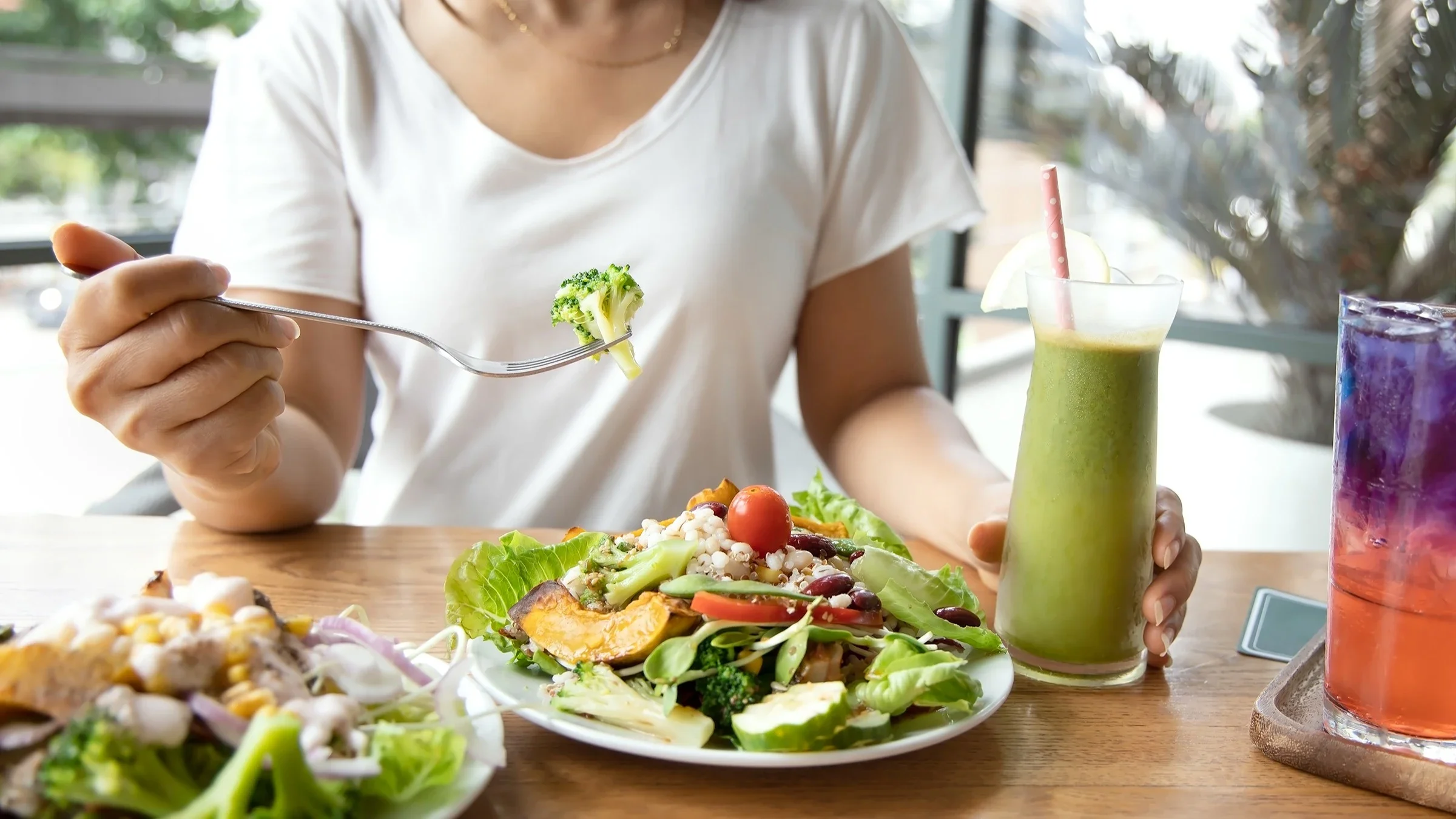Cropped shot of a person eating a large plate of veggies for lunch. They also have a green smoothie with a pink polkadot straw in it.
Sasithorn Phuapankasemsuk/iStock via Getty Images