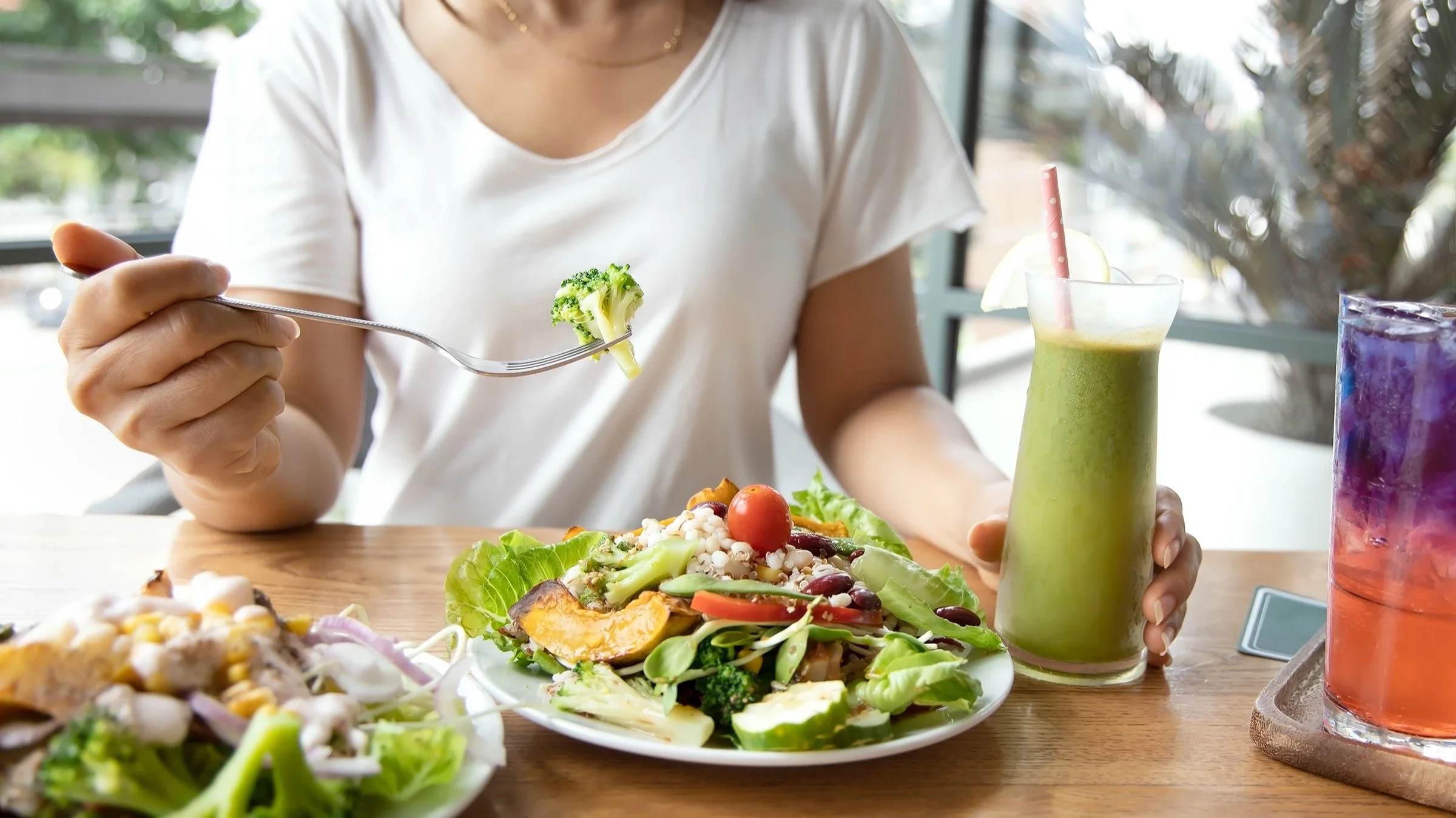 Cropped shot of a person eating a large plate of veggies for lunch. They also have a green smoothie with a pink polkadot straw in it.