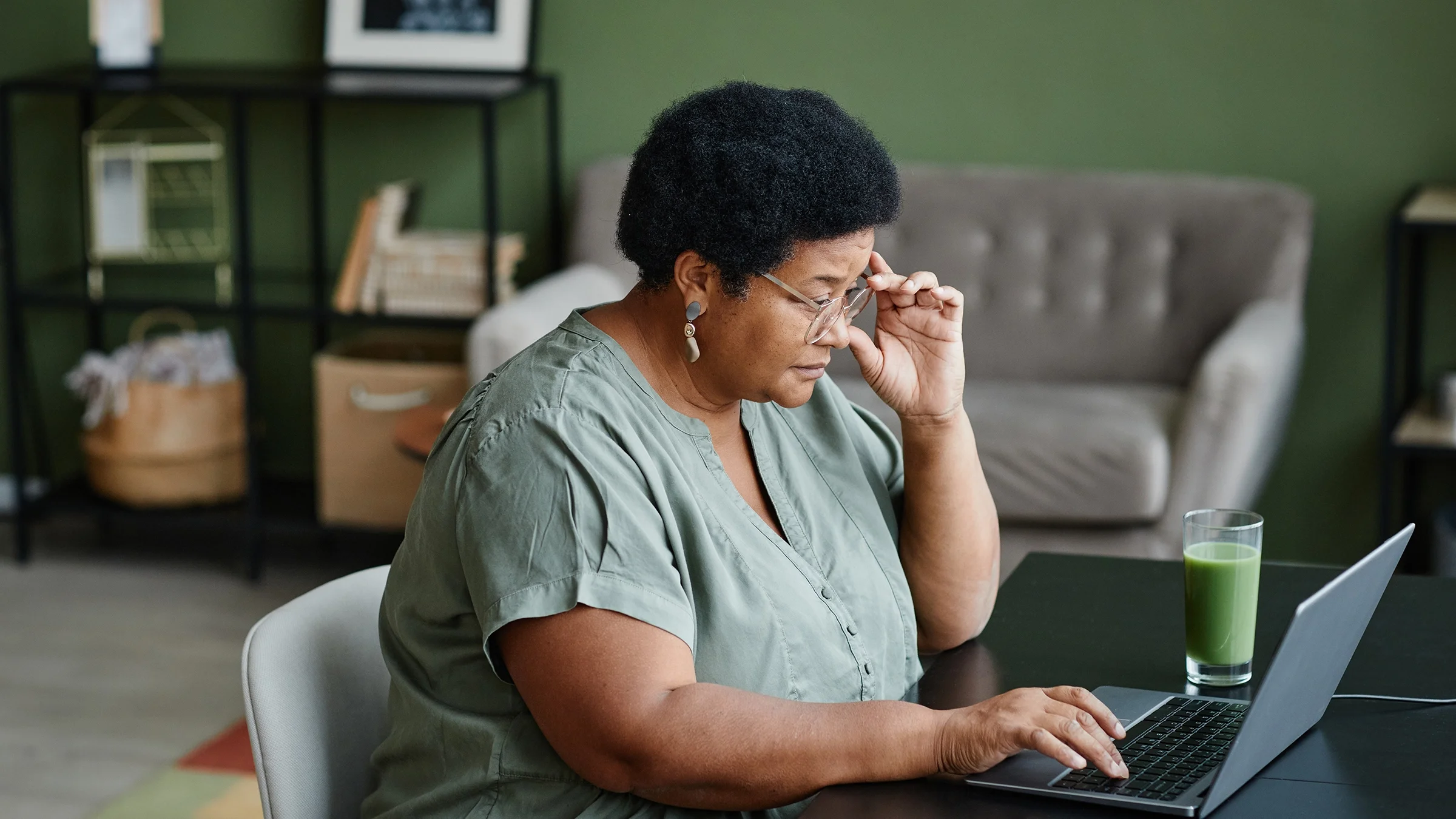 A woman sits at her kitchen table with her laptop and a green juice.