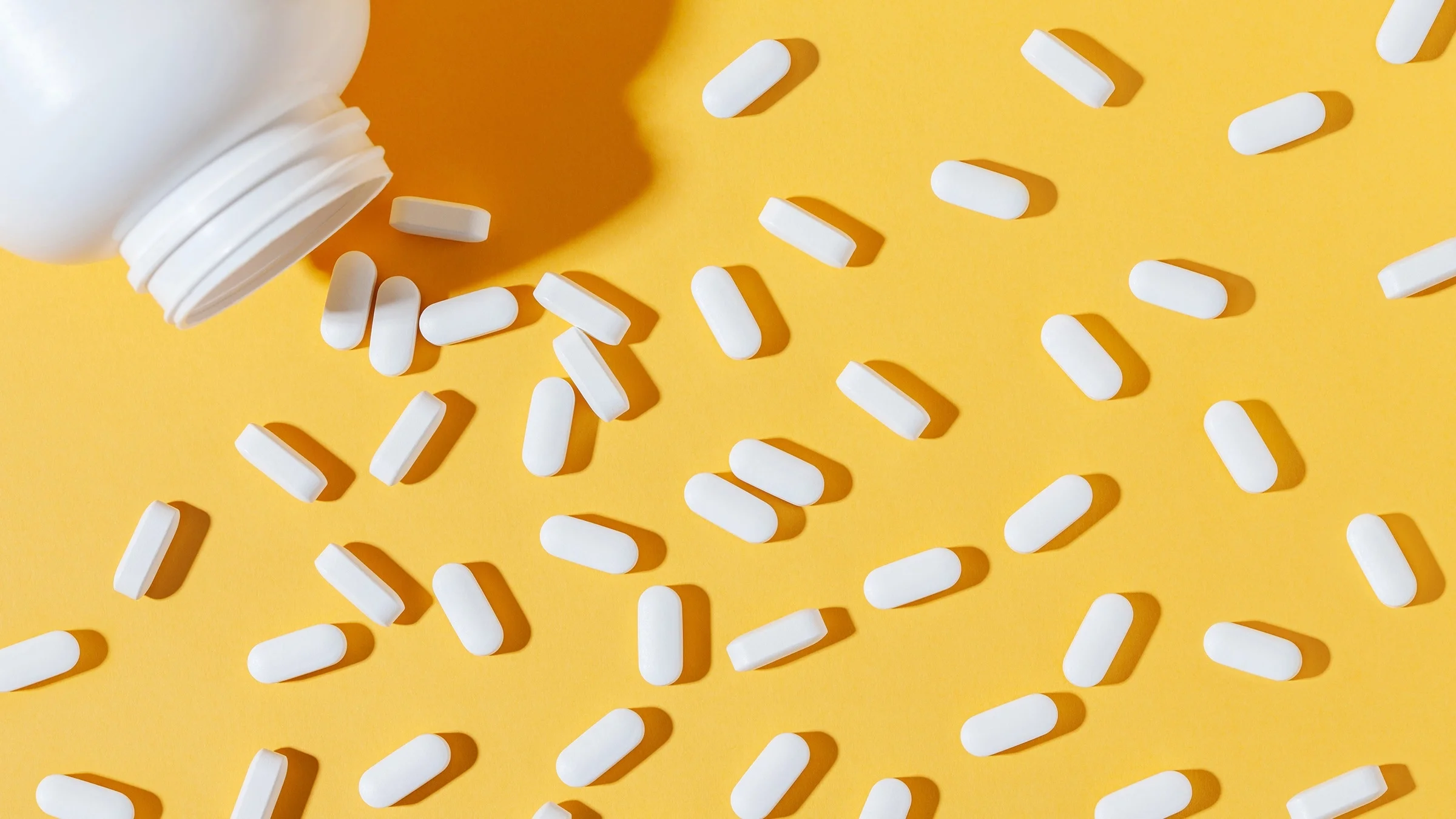 White pill bottle with white oblong pills spilled out across the frame on a yellow background.