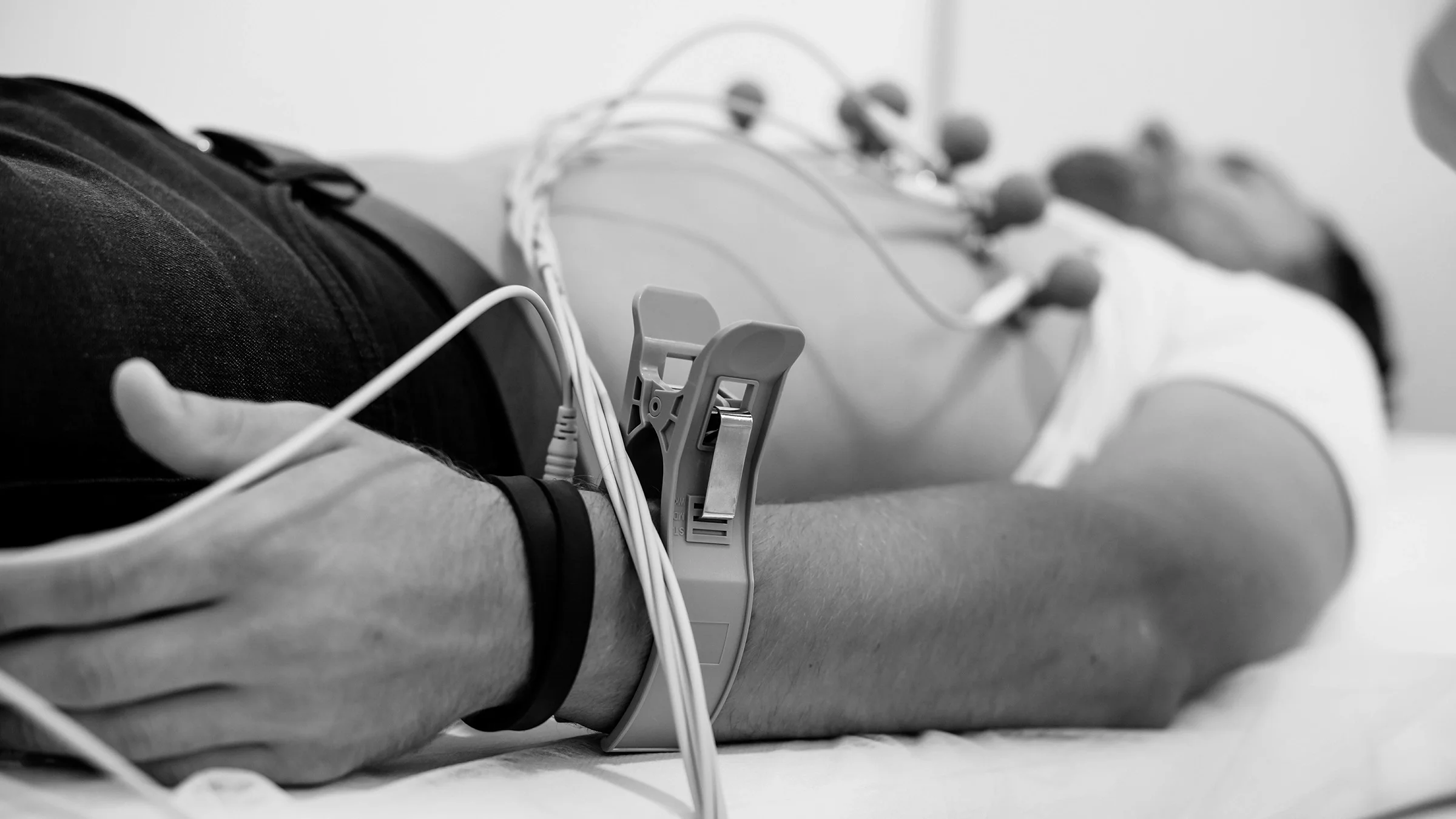 Black and white close-up of a man's wrist as he is lying down and attached to EKG equipment for a test.