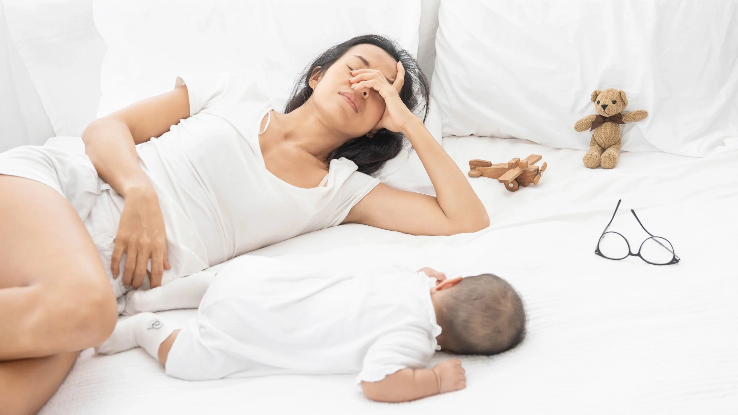 A tired mom laying in bed next to her baby and toys.