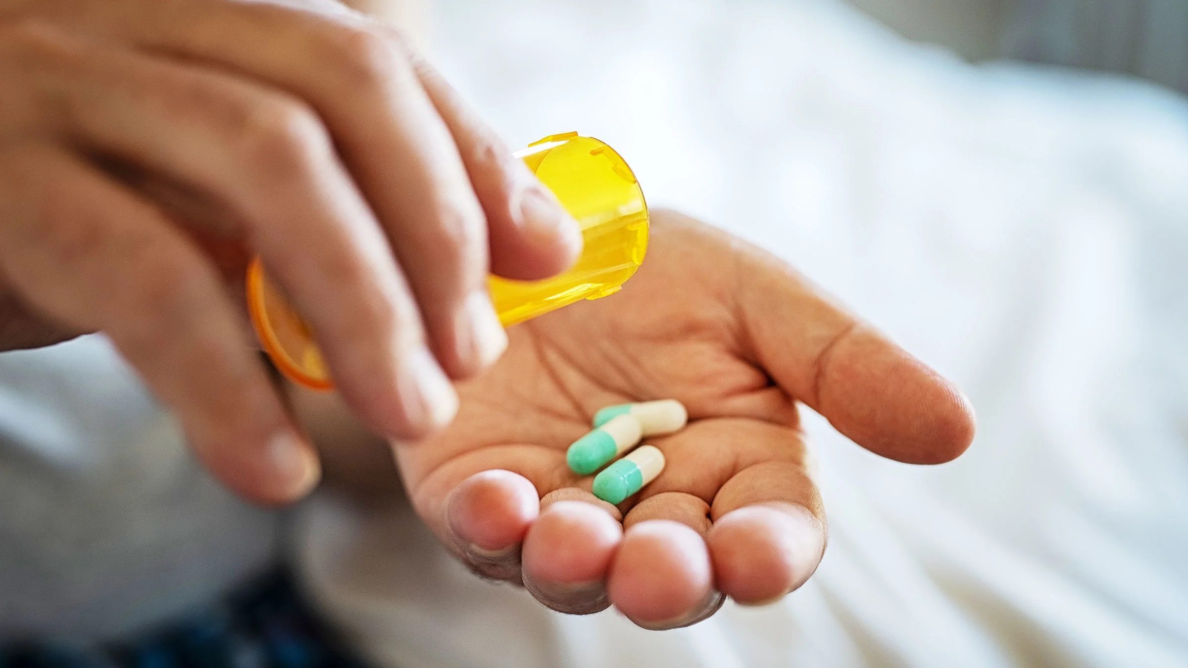 Close-up of a man pouring teal and white capsule pills into his hand from the yellow pill bottle.