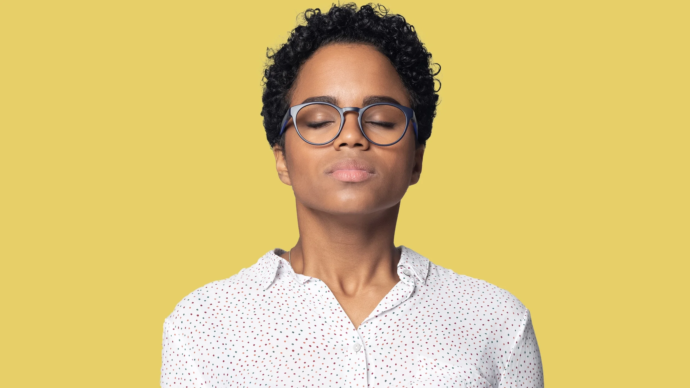 Studio portrait of a woman with her eyes closed and taking a deep breath in. She is on a plain yellow background.