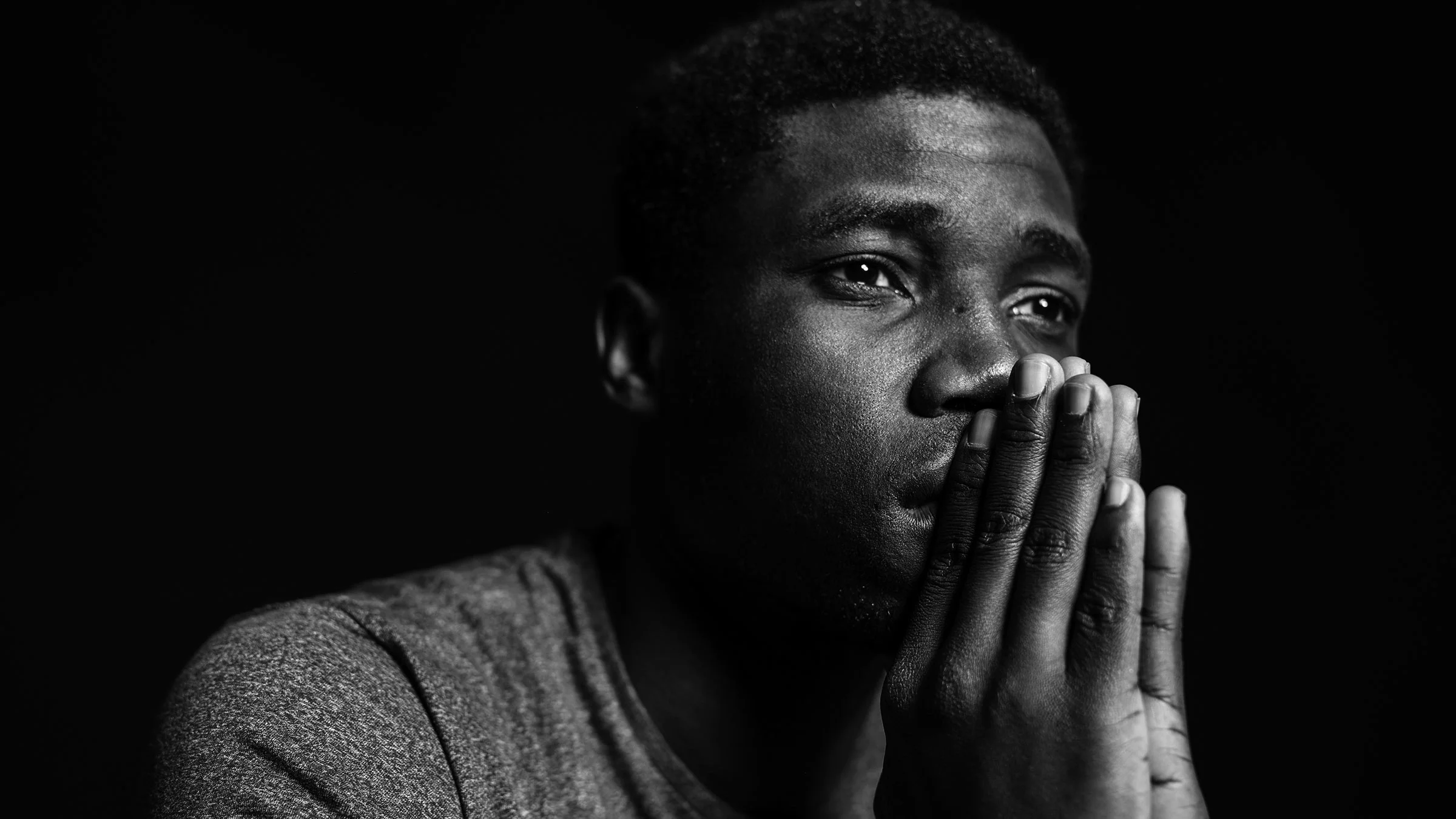 Black and white close-up of a man with his hands clasped together in front of his face on a plain background.