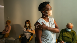 Woman standing in a clinic holding a cotton swab on her arm where she just got a vaccine shot. There are other's sitting behind her in the waiting room.
Dimensions/E+ via Getty Images