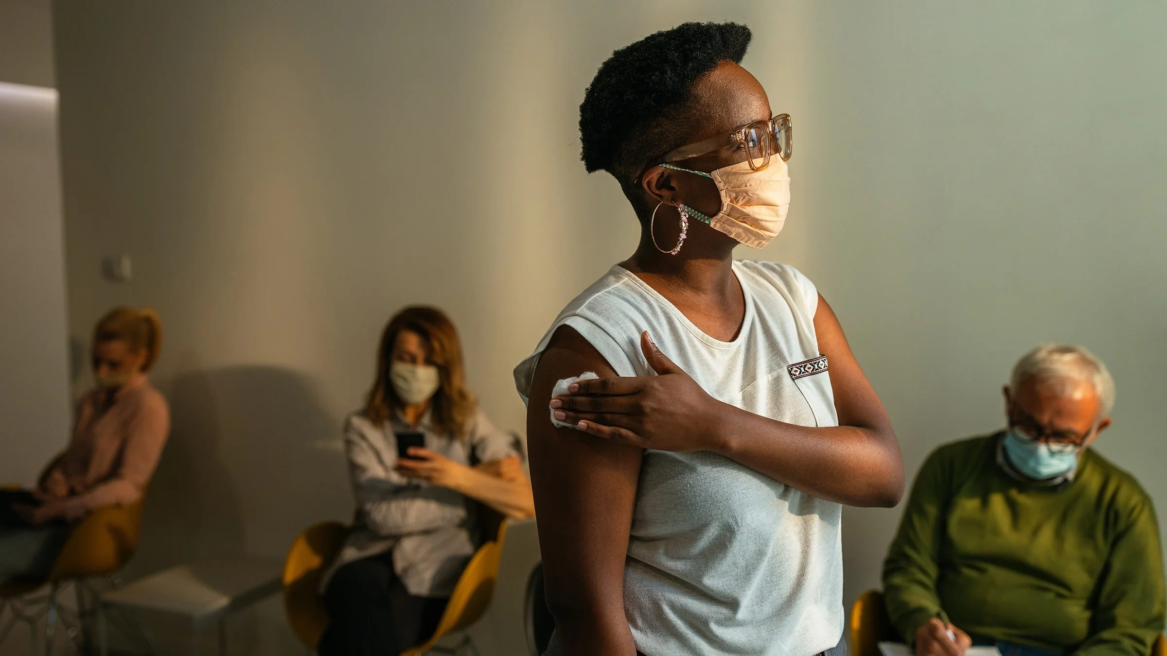 Woman standing in a clinic holding a cotton swab on her arm where she just got a vaccine shot. There are other's sitting behind her in the waiting room.