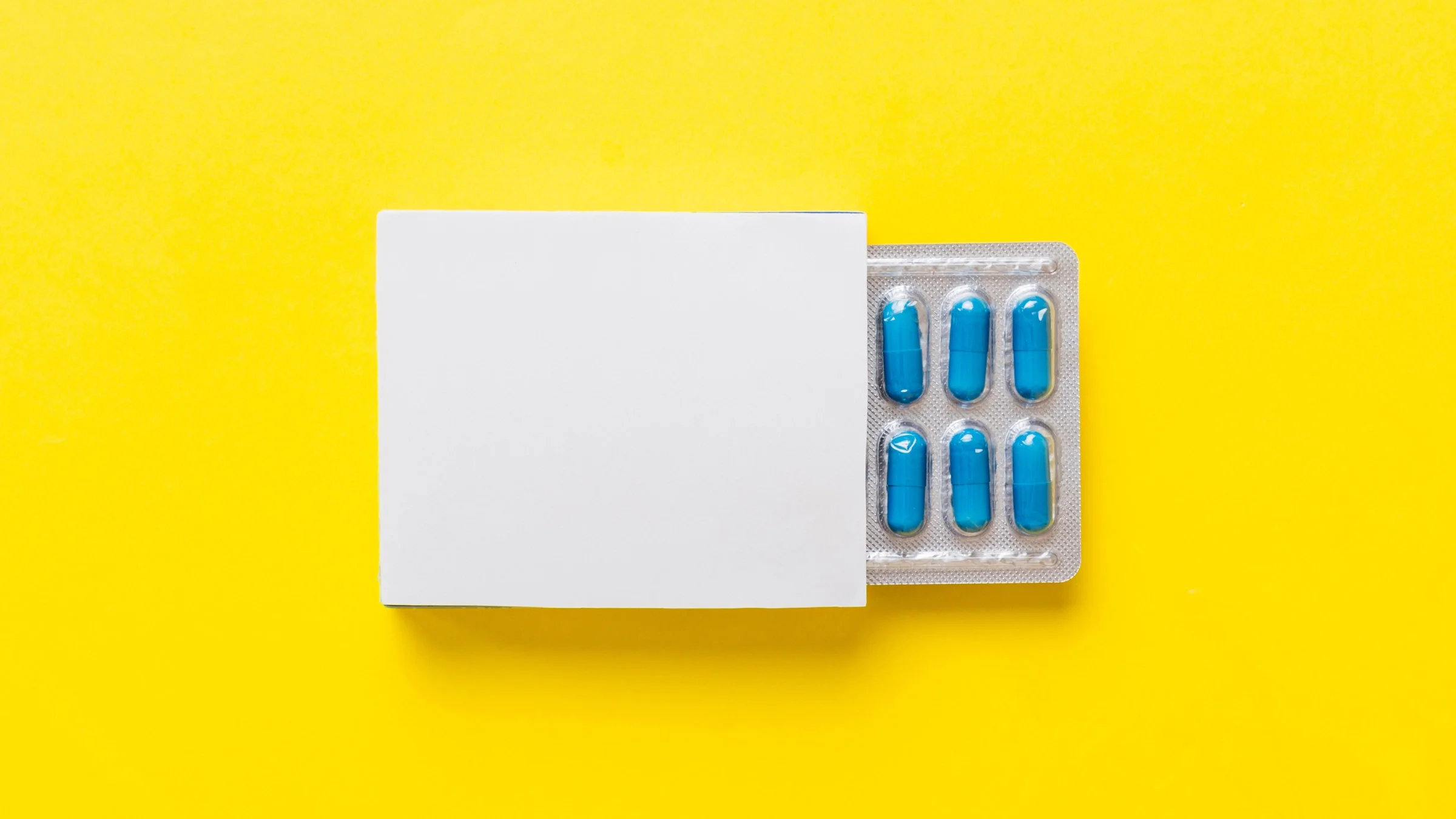 Blister pack of blue capsule pills coming out of a white medication box on a bright yellow background.