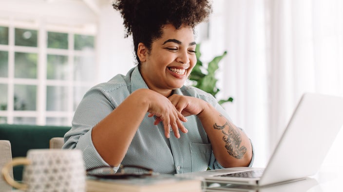 Young woman with curly hair up high on her head smiling big as she talks to someone on video chat on her laptop.