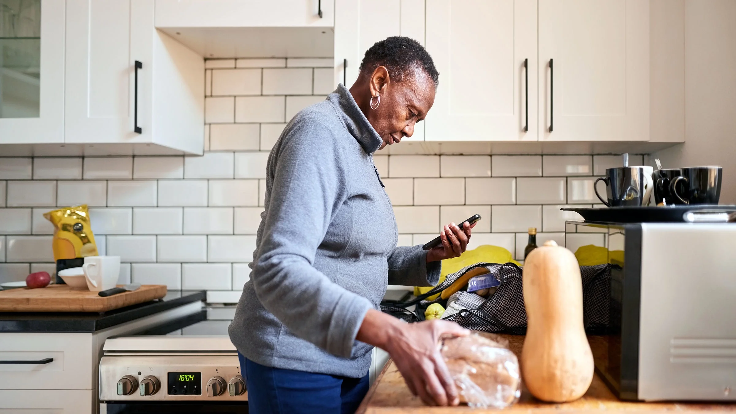 A senior woman uses a mobile phone while unpacking groceries in the kitchen.
