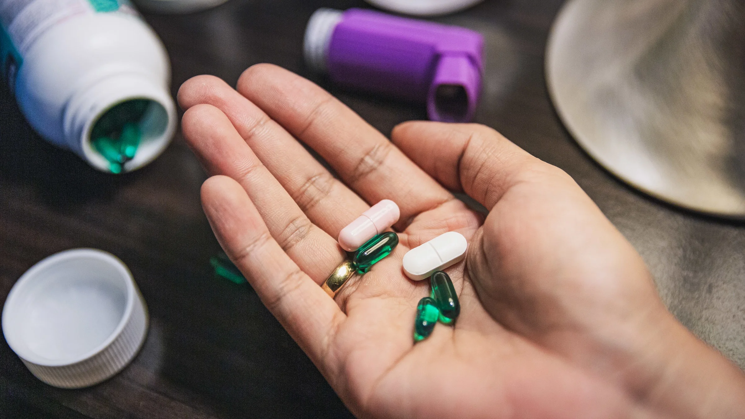 Close-up of a woman's palm with various pills in hand, inhaler and pill bottle on the table as well.
