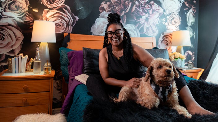 Nikki Nurse at home in bed with her dog, Nino. They are in front of a wallpapered wall with oversized roses. 