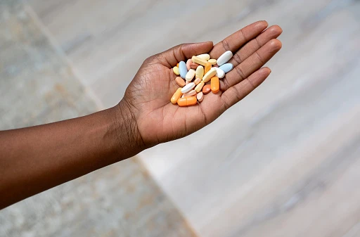 Close-up on a a woman's hand holding a palmful of assorted orange, pink, white, yellow, and blue pills on a textured stone background.