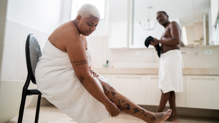 A woman shaves her legs in the bathroom while sitting down in a chair.