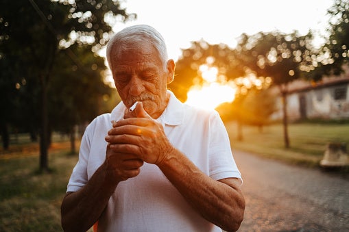 Older man smoking a cigarette outside during sunset.