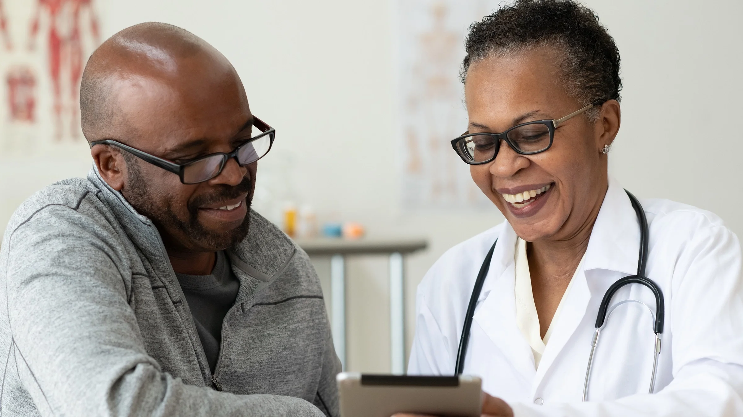 Older Black man reviewing his medical chart with his doctor on a tablet device. They are sitting in the exam room next to each other and they are both wearing black plastic glasses.