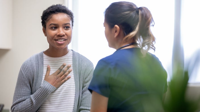 Young female patient with her hand on her chest talking to a nurse in an exam room with a slight smile.