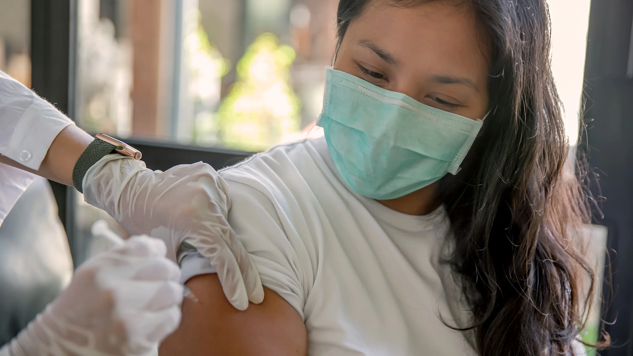Close-up of a woman getting a shot. She is wearing a green-blue medical face mask. The nurse administering the shot is wearing latex gloves.