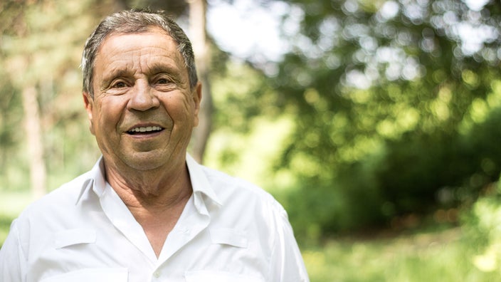 Portrait of an elderly man standing outside in a park. There is a lush green background of trees behind him.
