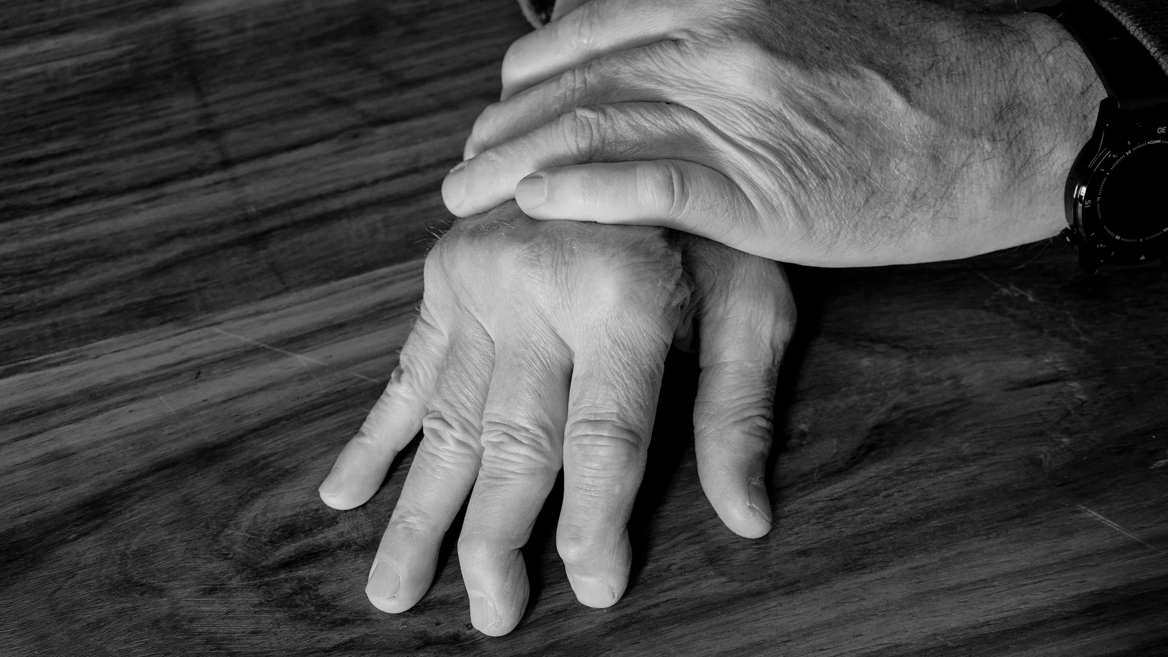 Black and white close-up of a man's hands crossed. He has arthritis in his fingers.