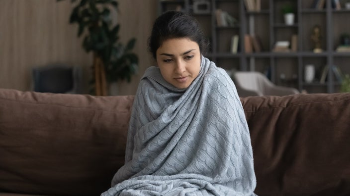 Young woman sitting on the couch wrapped up in a blanket to her neck, with only her head exposed.