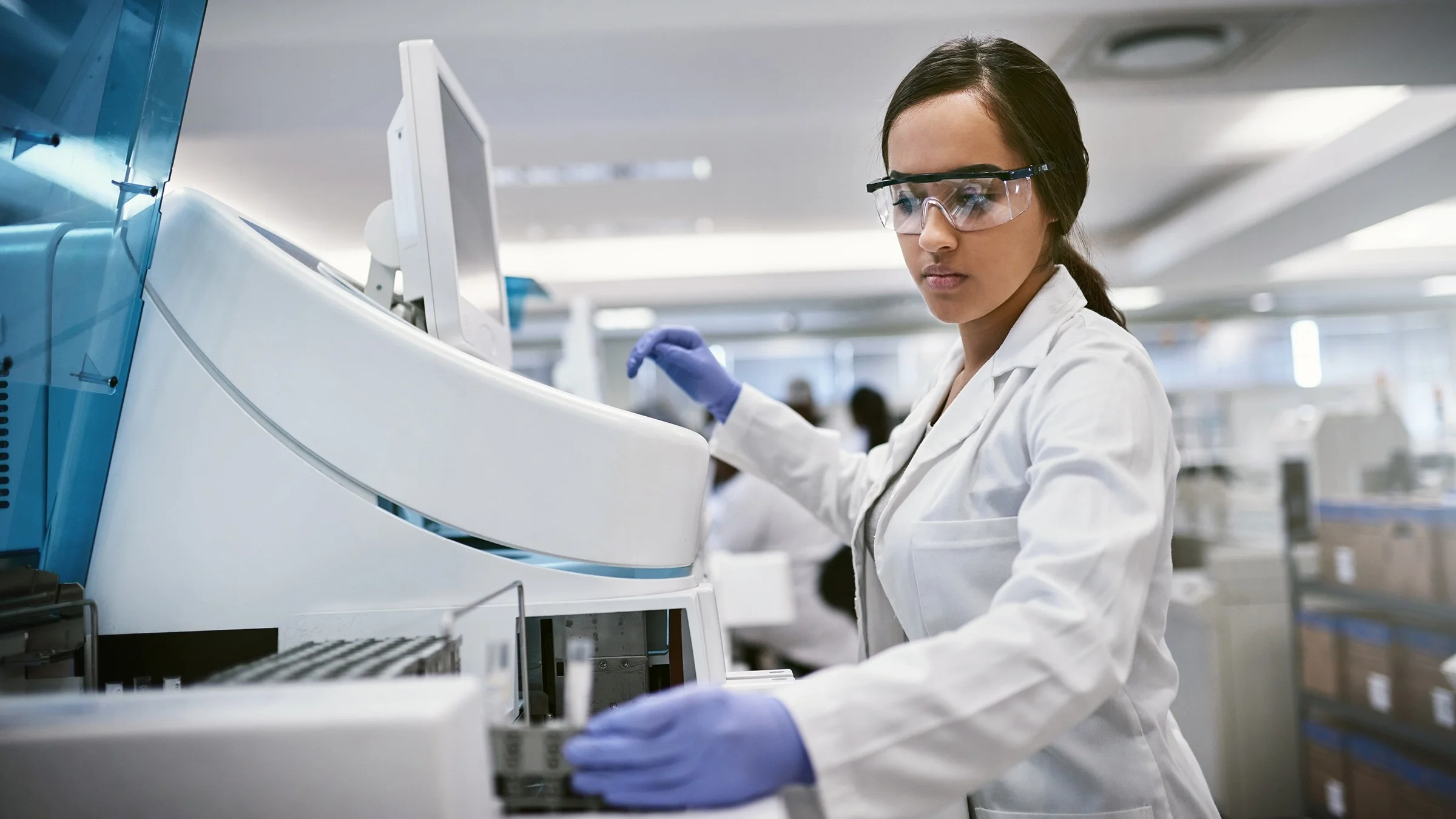 Young lab technician using a large machine in a laboratory to process samples. She has her hair tied back and is wearing protective glasses and blue medical gloves.