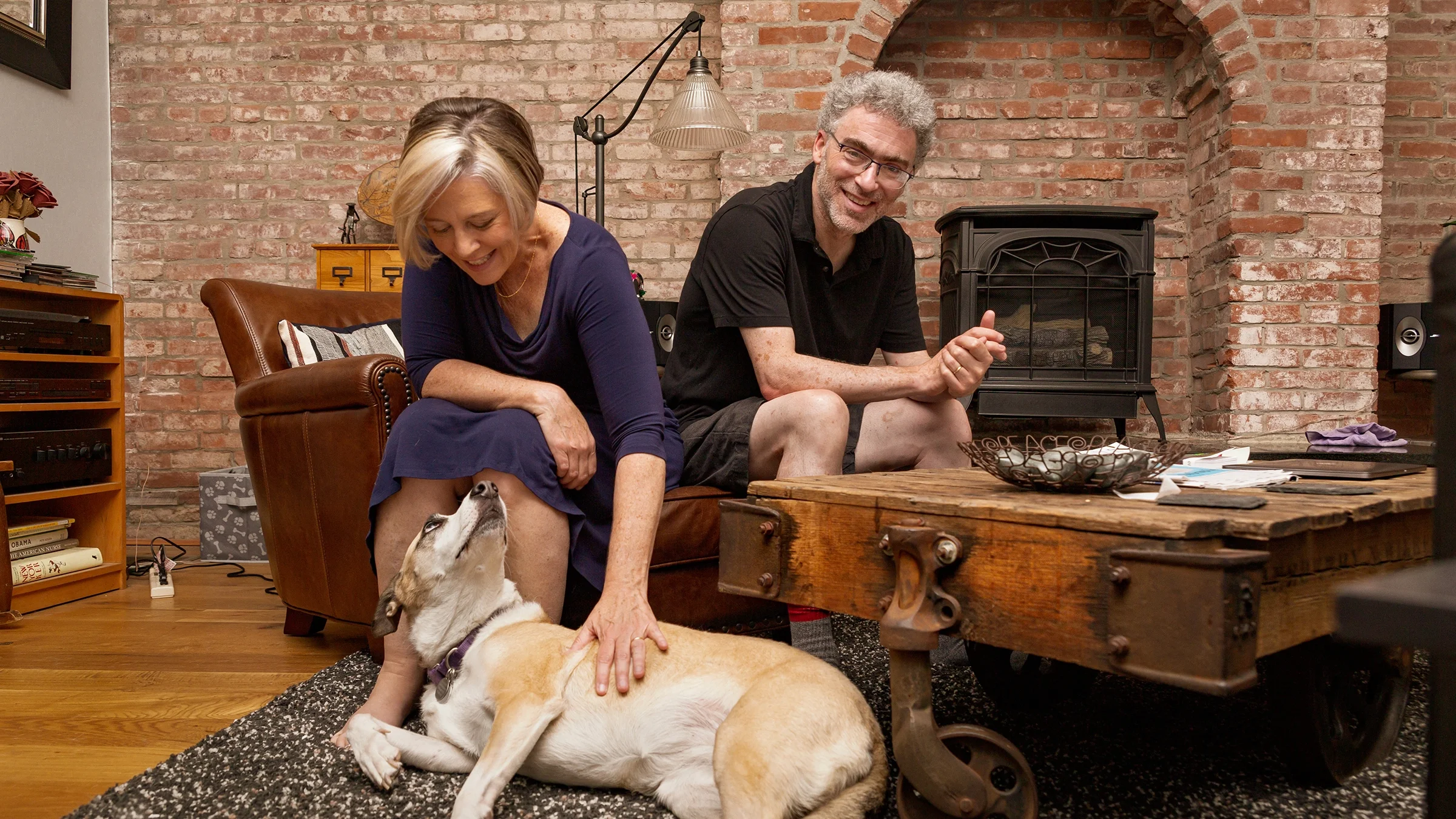 Breast cancer survivorTheresa Brown at home with her husband, reaching down to pet her dog as they sit by the fireplace.