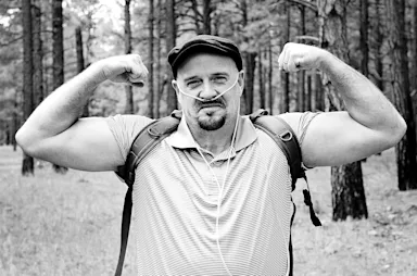 Black and white portrait of a man walking in the forest with nasal cannula oxygen on and flexing his biceps.
carolthacker/iStock via Getty Images