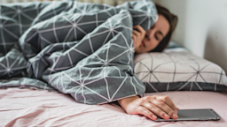 Woman covered in a gray comforter reaches for her phone in bed.
blackCAT/E+ via Getty Images