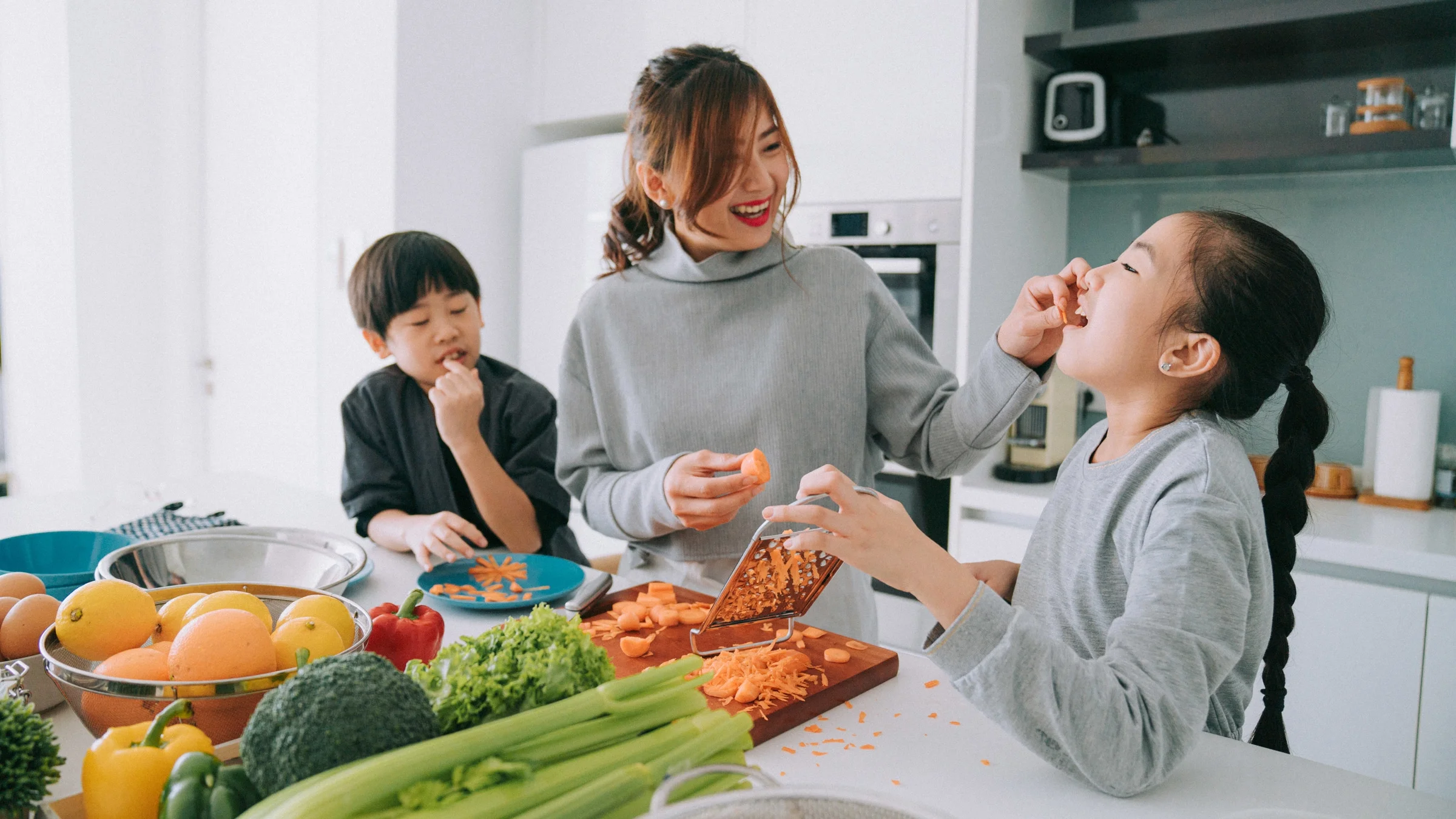 Smiling mother feeding her daughter a string of carrot, while her brother watches on the left. They are in the process of shredding a carrot together, and there are other fruits and vegetables on the table.