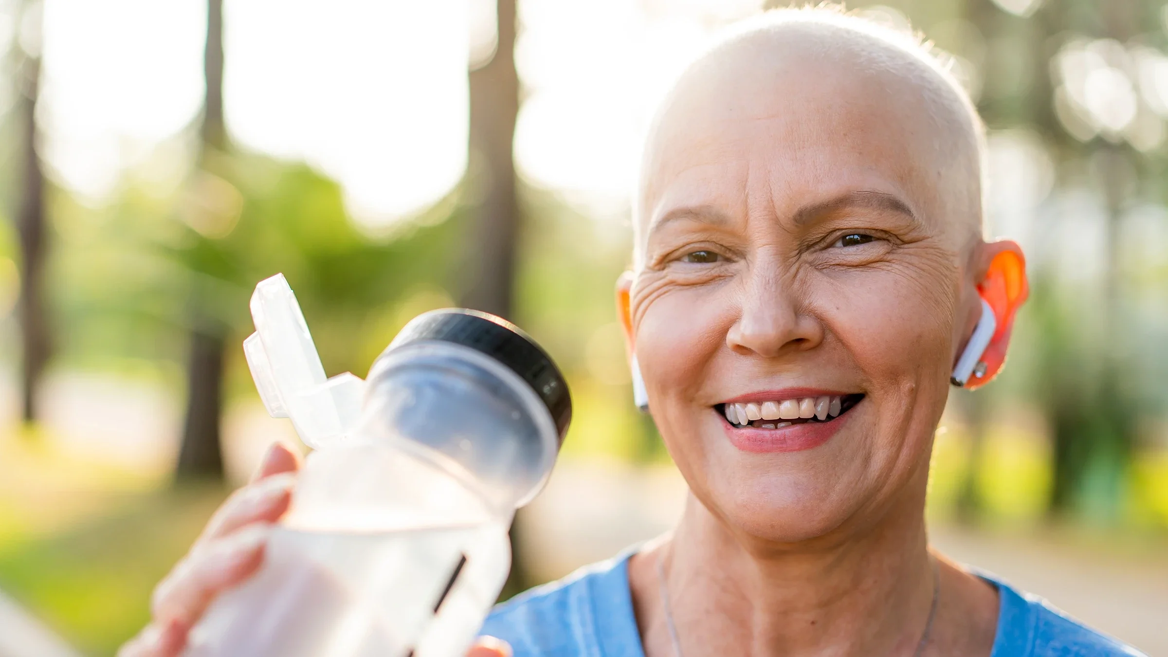Portrait of a woman smiling with ear buds and drinking water.