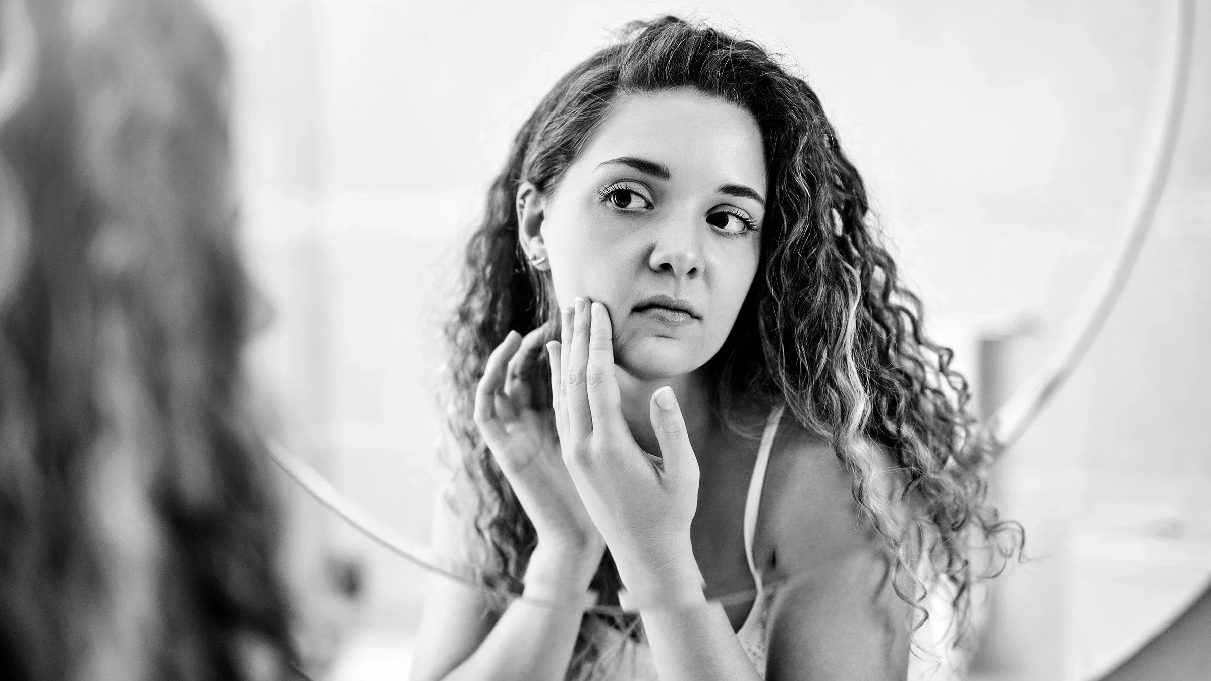 Black-and-white image of a woman checking her chin for pimples in the mirror.
Charday Penn/iStock via Getty Images