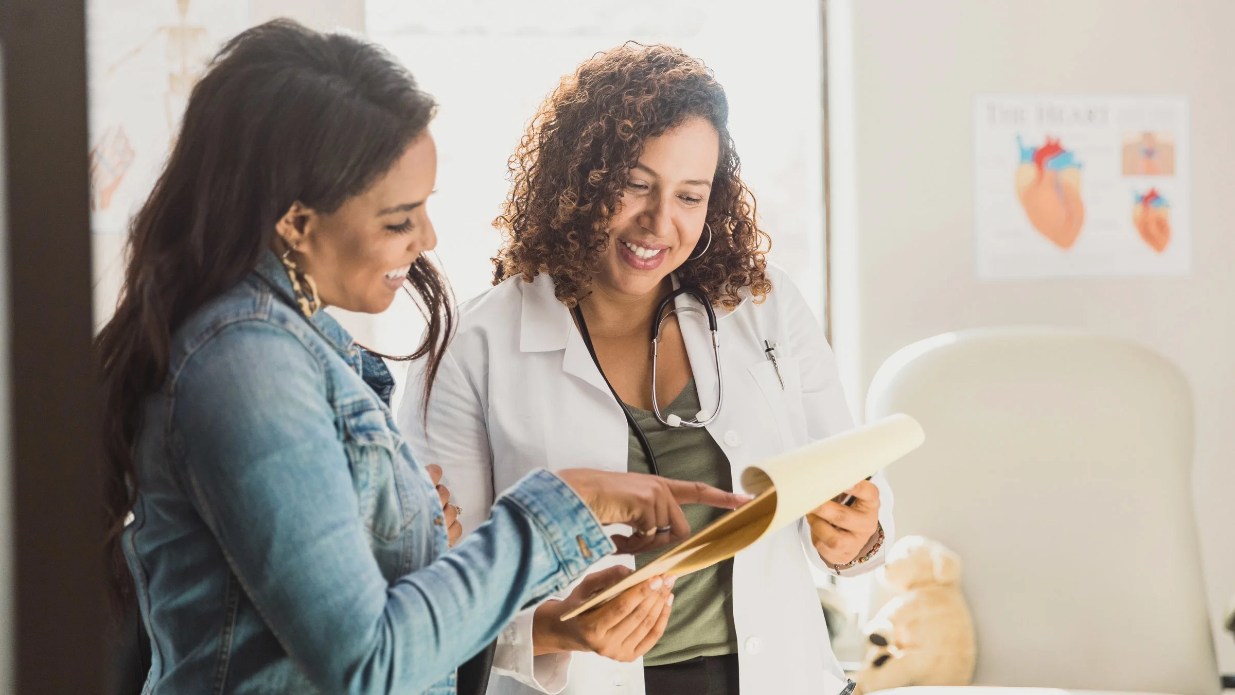 Female gynecologist and  female patient both smile as they review medical paperwork on clipboard in exam room.