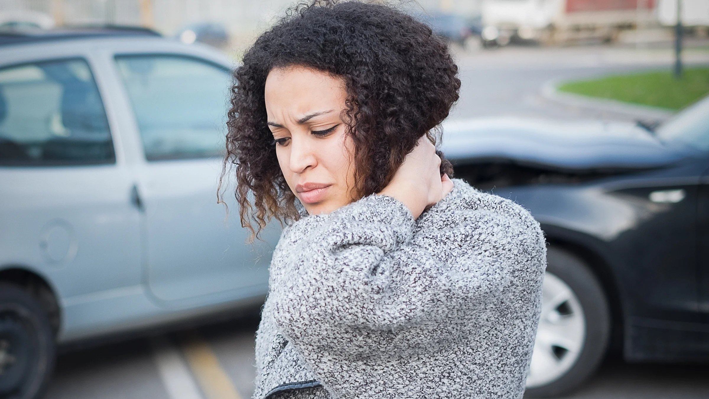 A woman is holding her neck after a whiplash injury in a car accident, which is visible in the background behind her.