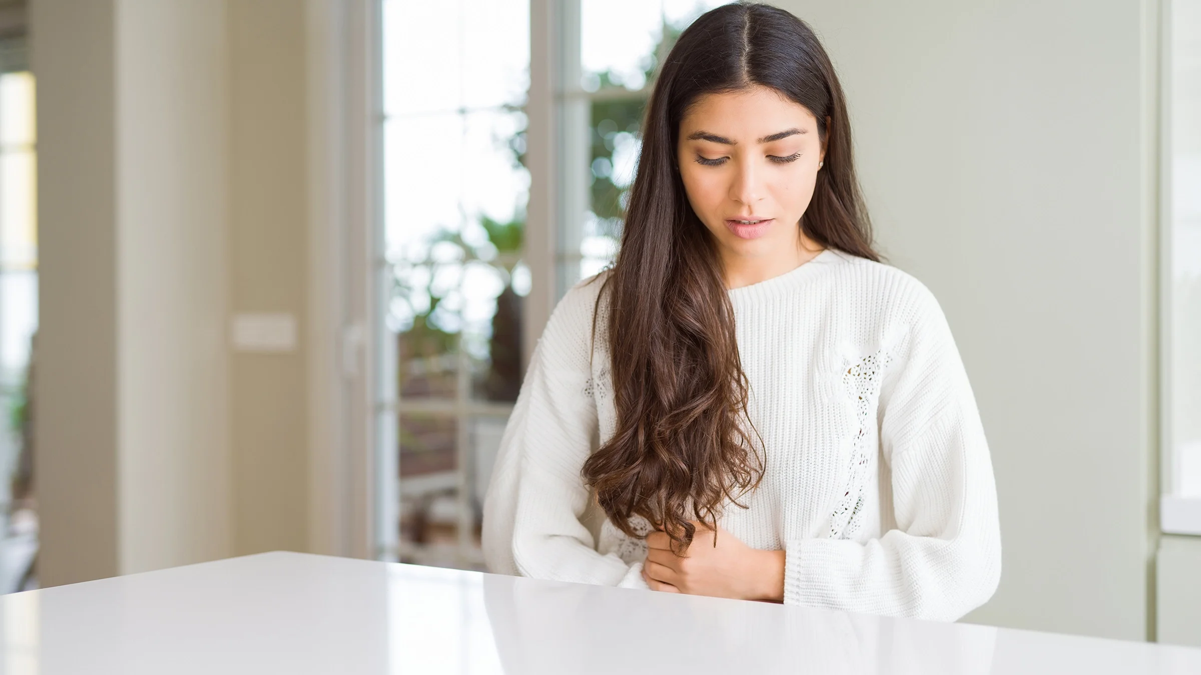 Woman with a stomach ache grabbing her stomach as she stands in the kitchen.