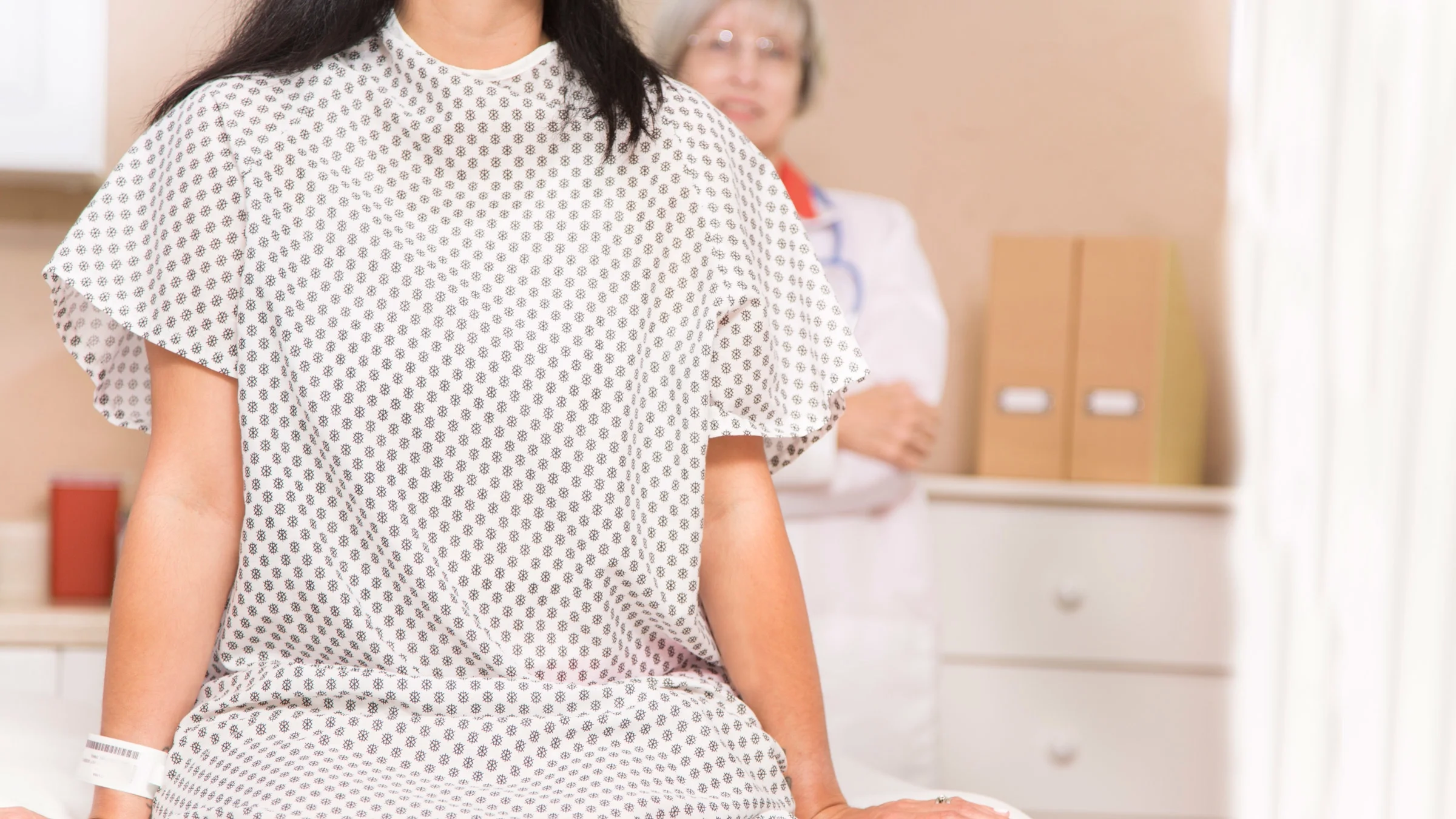 Cropped shot of someone sitting on a surgical table in a hospital gown. Their doctor is standing behind them.