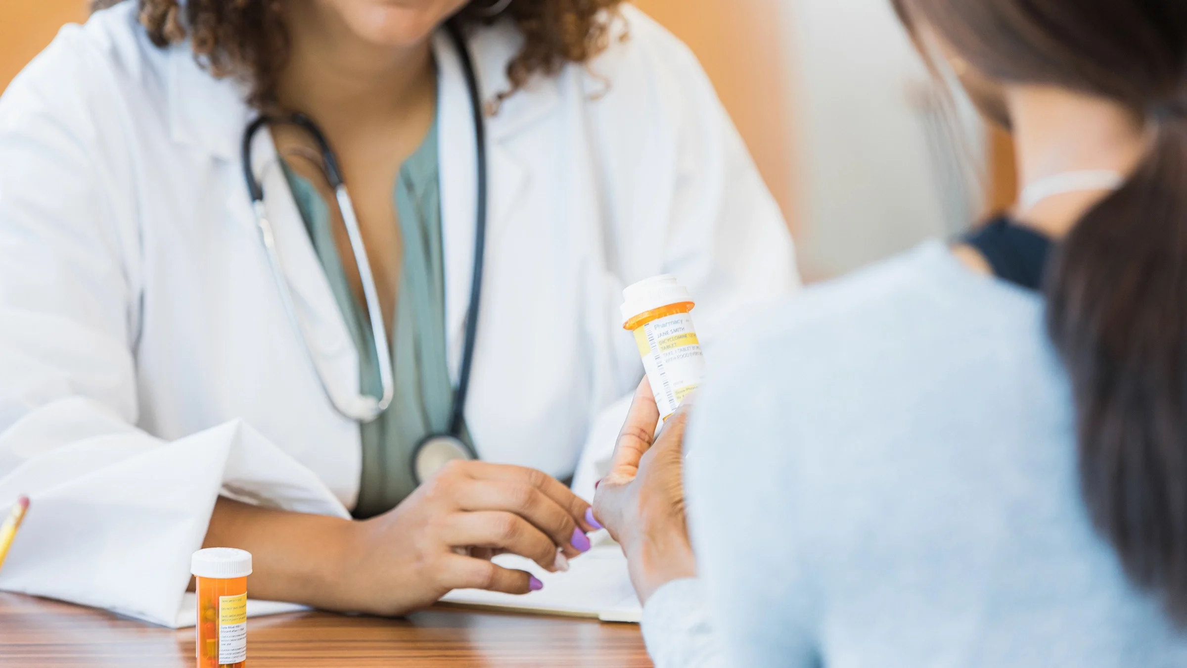 Doctor reviewing a prescription with the patient. The image is cropped in a way where we can't see either person's face. The focus is on the orange prescription bottle in the middle.