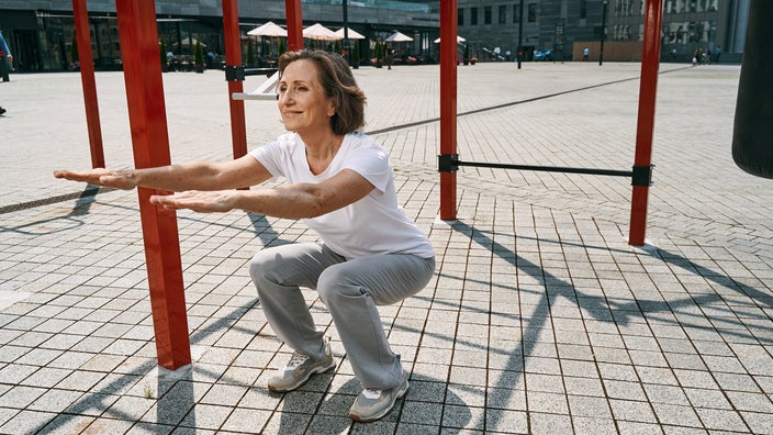A woman is doing squat exercises while training on a sunny day in a city square.
