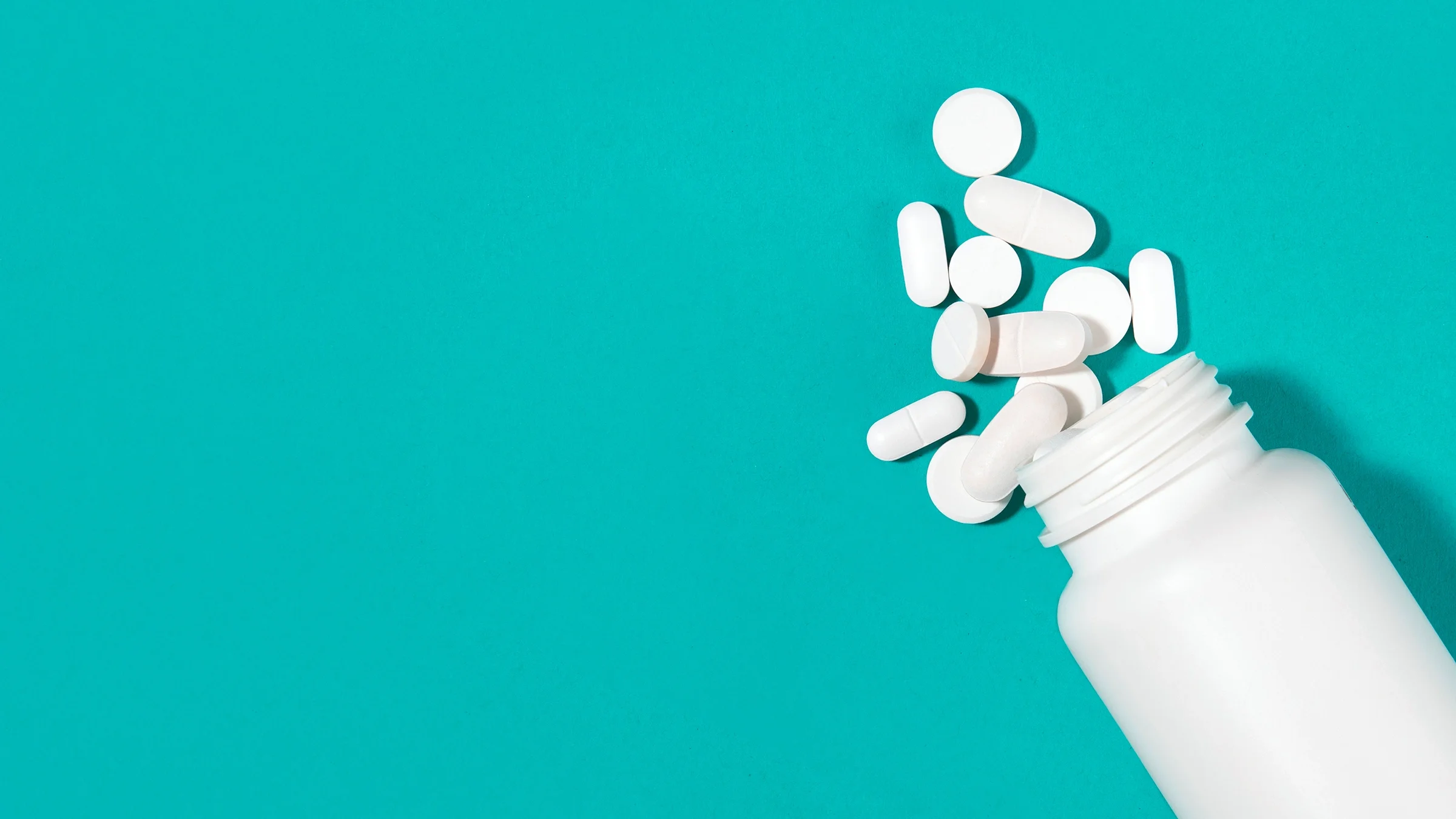 White medicine bottle with white pills spilling out on a teal background.