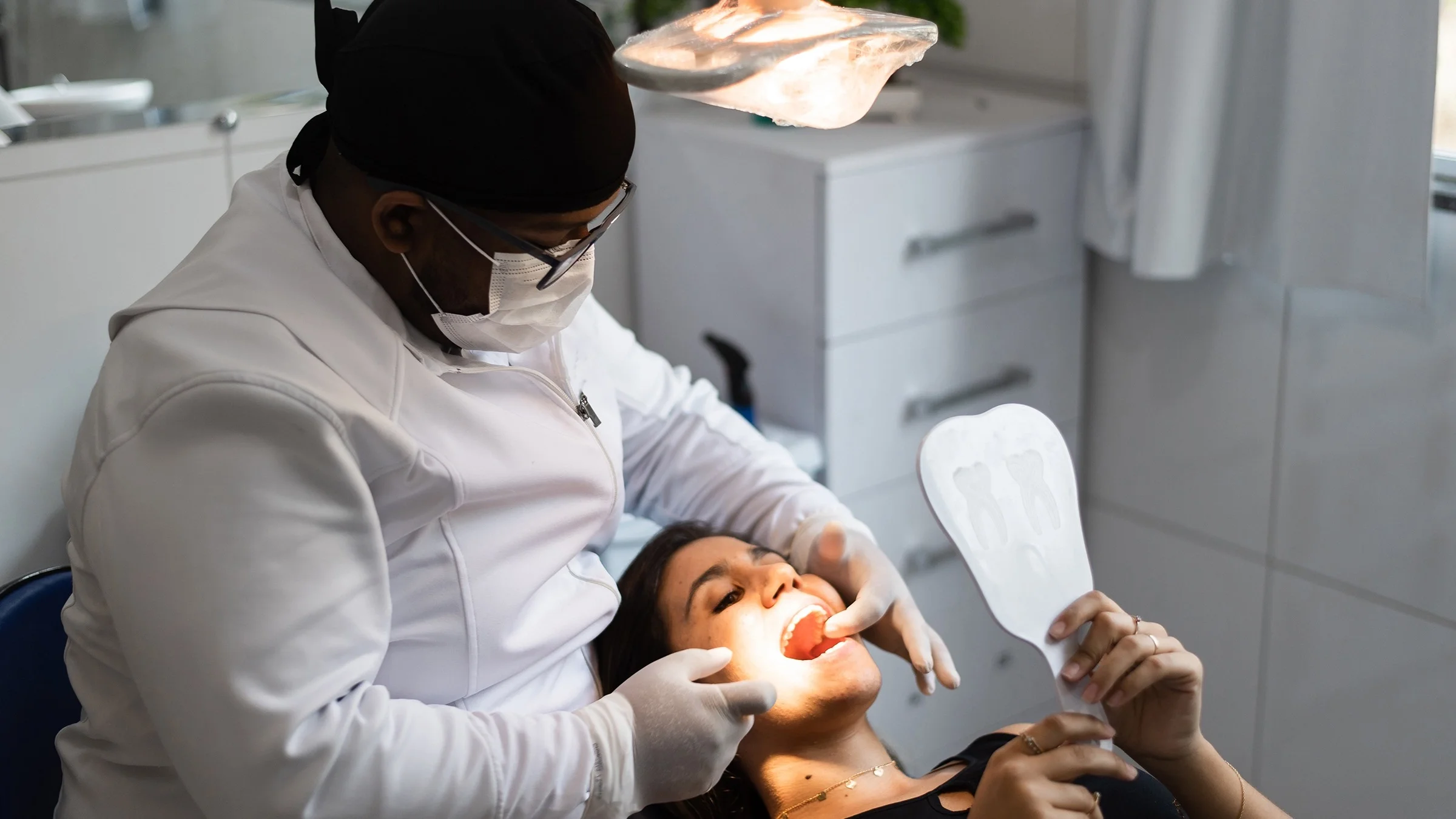 Young woman in the dentist chair getting her check up. The dentist is pointing out problem areas for her as she uses a hand-held mirror to see in her mouth.