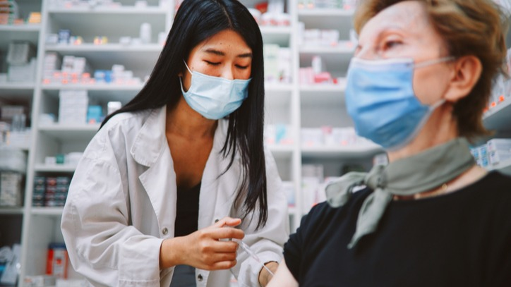 Elderly woman receiving a vaccine at the pharmacy, both pharmacist and patient are wearing blue medical face masks.